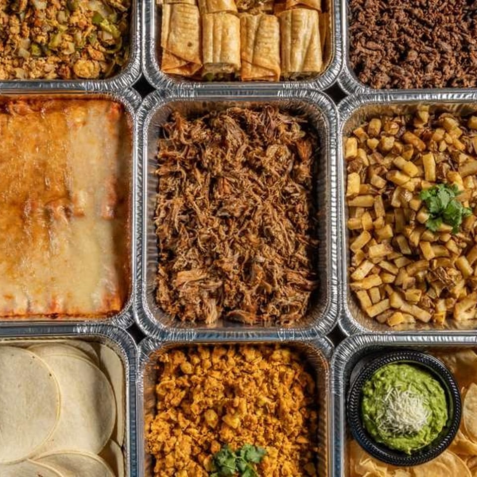 Assorted Mexican dishes, including shredded meats, rice, beans, tortillas, enchiladas, potatoes, guacamole, and chips, arranged in foil trays.