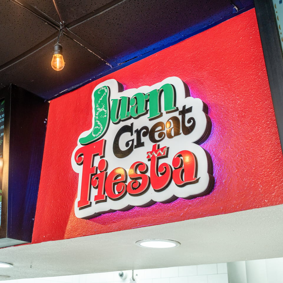 Colorful Juan Great Fiesta sign mounted on a red wall inside a Mexican restaurant, with menu boards and hanging lights.